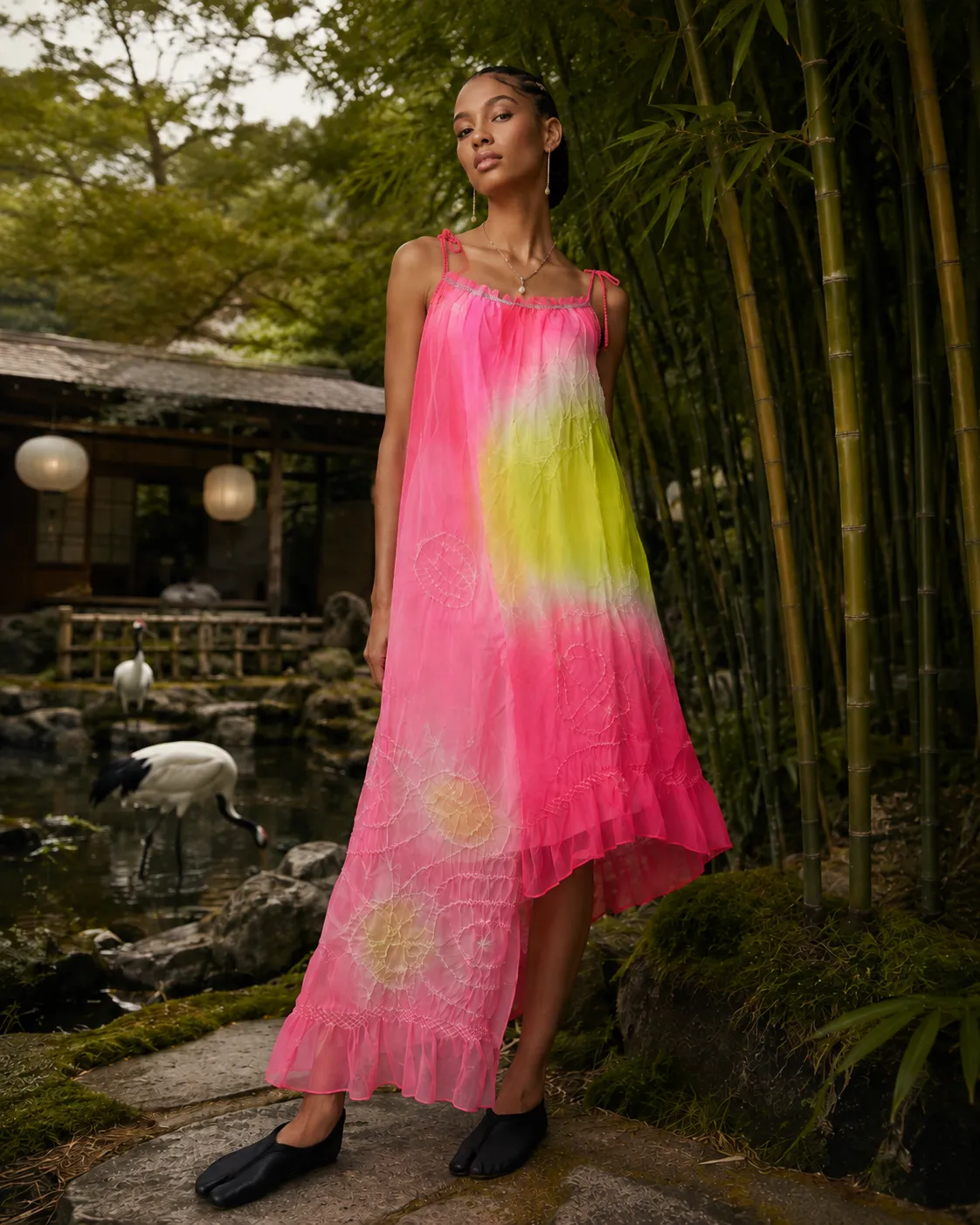 AI-developed campaign image: a model in a long pink and yellow tie-dyed dress, standing in a Japanese garden with bamboo and a traditional pavilion behind.