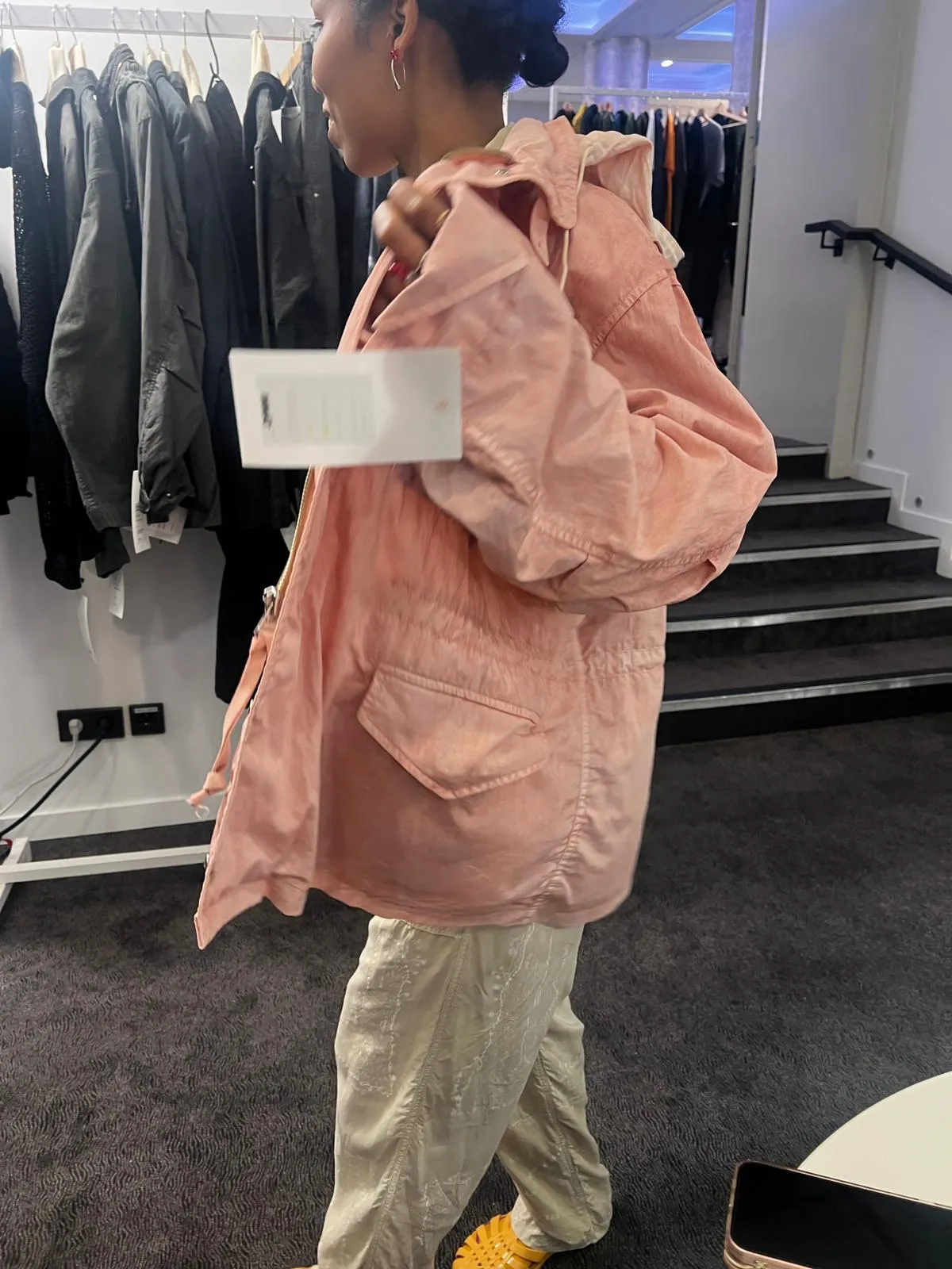An oversized pink jacket worn in profile on the showroom floor, in front of a rail of dark pieces.