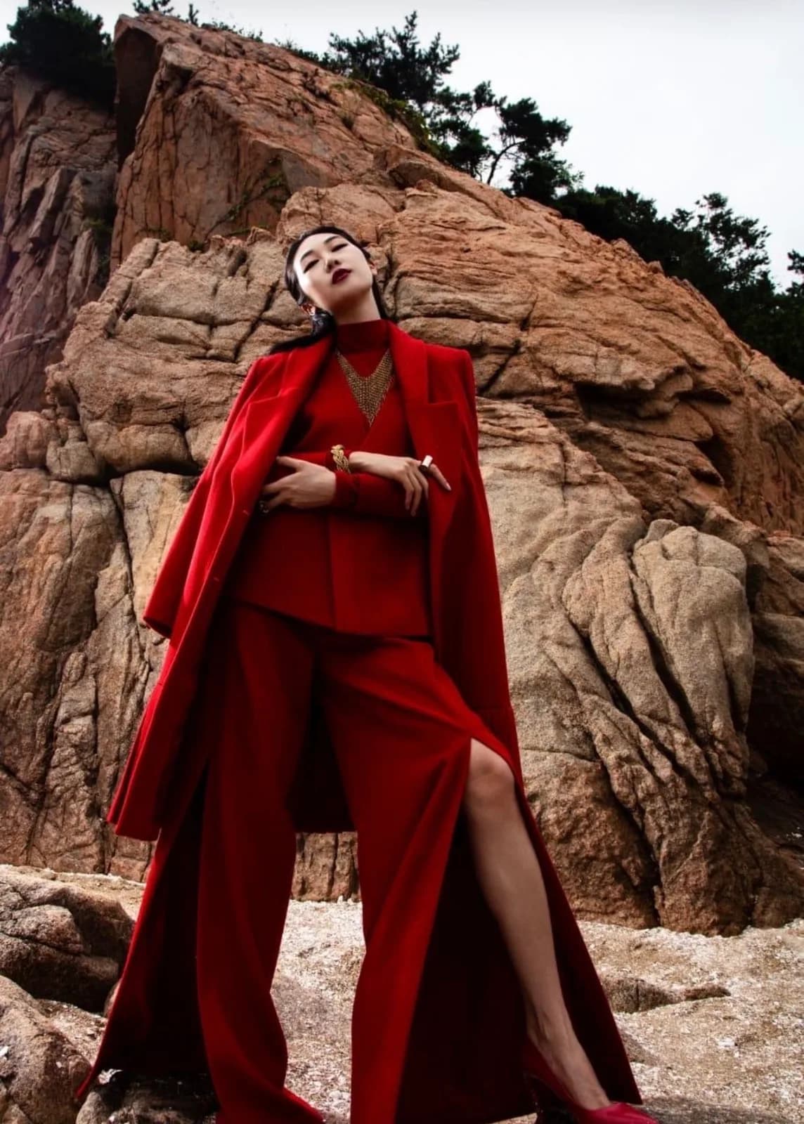 Editorial fashion photograph: a model in a full red look — tailored suit with a long cape coat — standing on rocks below a cliff face.
