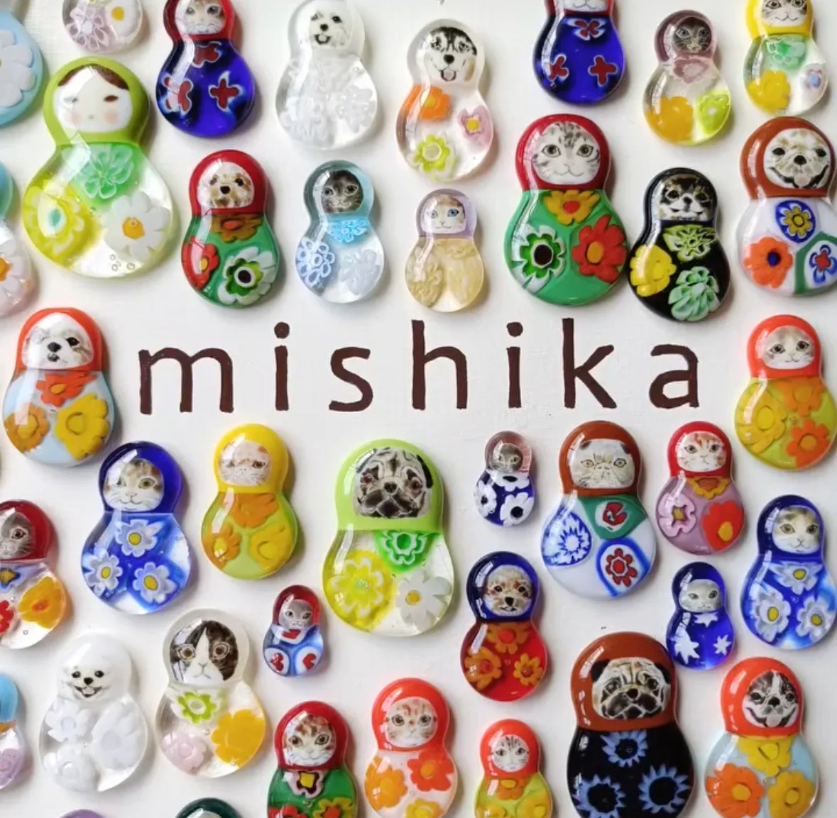 Mishika's handcrafted glass jewellery: dozens of matryoshka-shaped pieces in coloured glass, decorated with cats, dogs and flowers, arranged around the brand name.