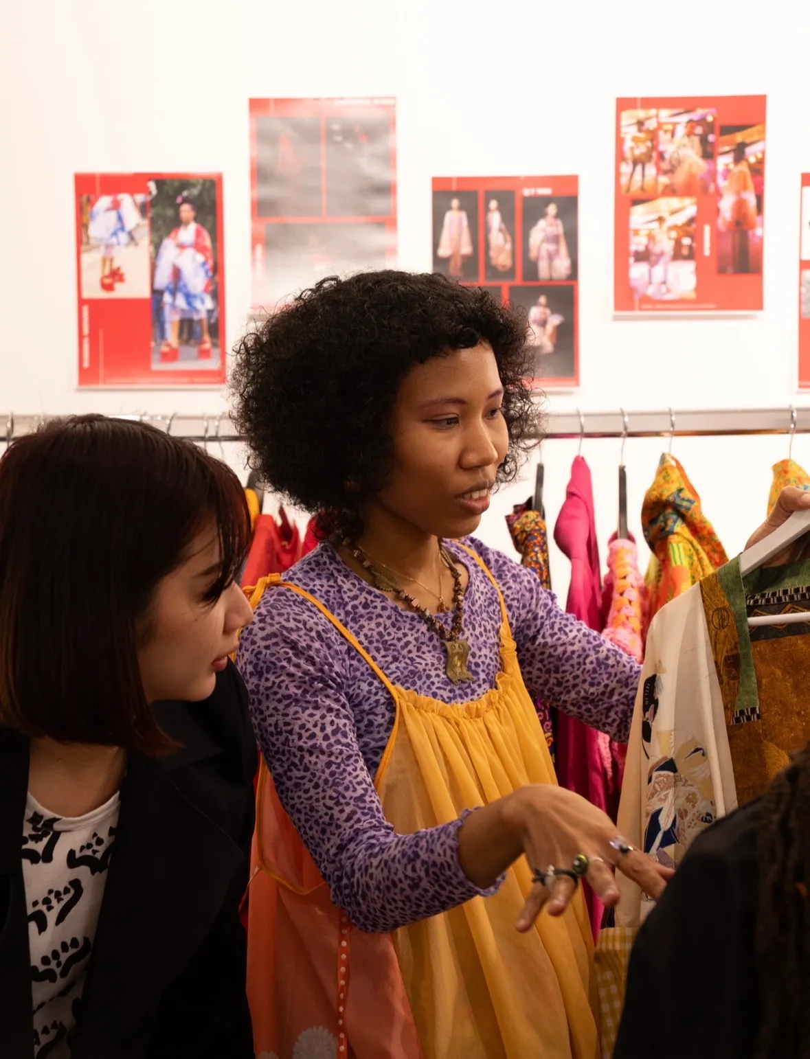 A woman in a purple leopard-print top and orange apron dress presenting a Tanowa Tomiyama garment on a hanger to a visitor, the collection on a rail behind her.